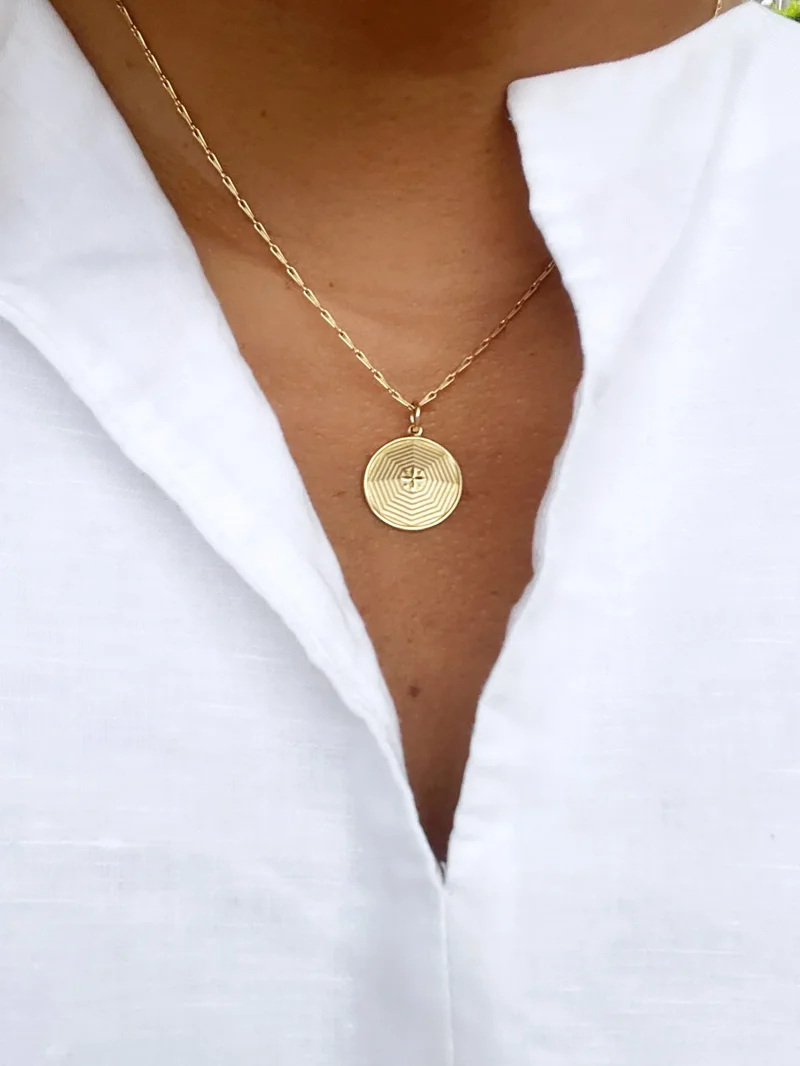 Gold circular pendant necklace with concentric line pattern and small cross detail, worn on model in white shirt