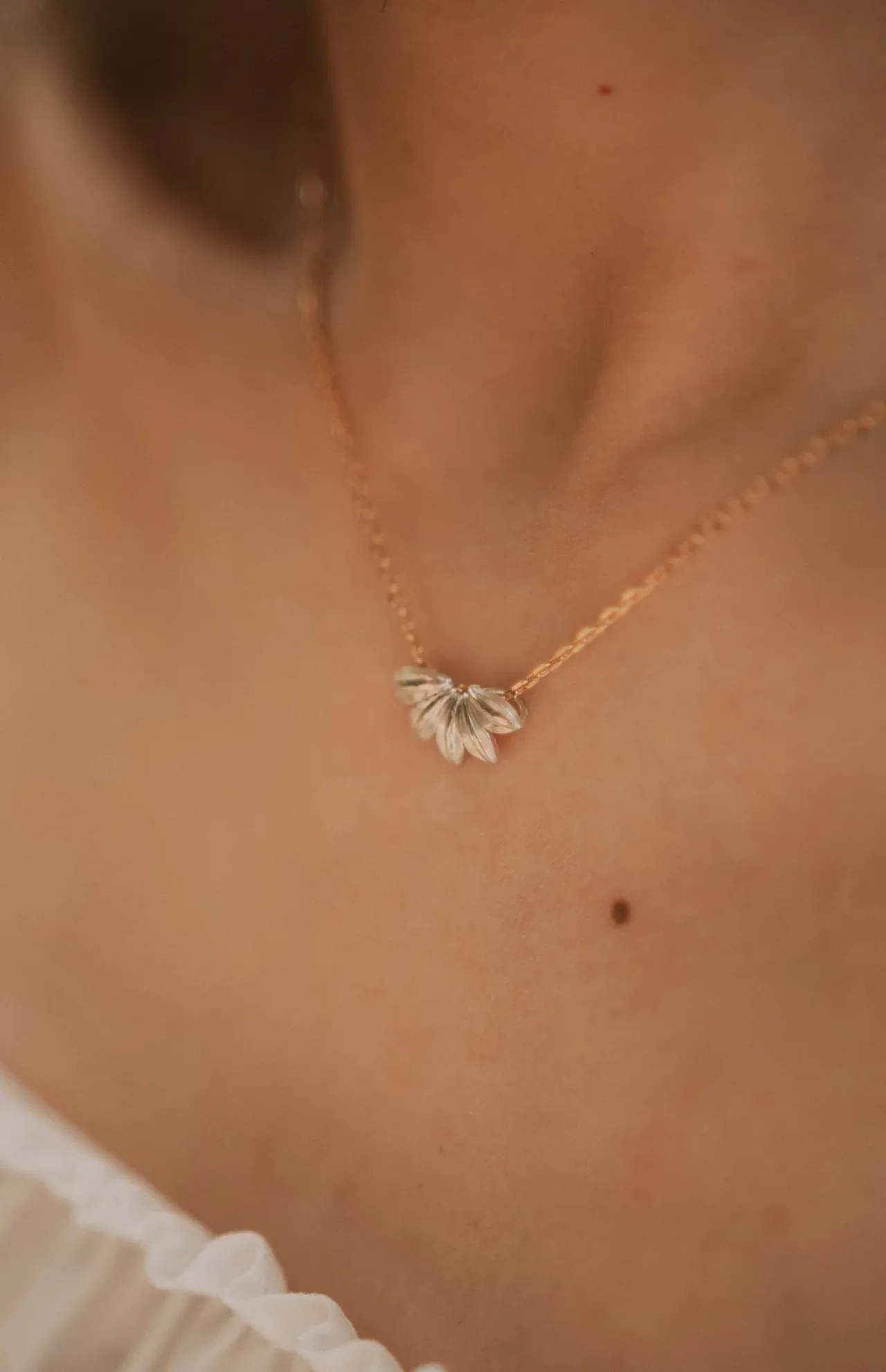 A delicate gold necklace with a small floral pendant rests on a person's neck, above a white ruffled garment.