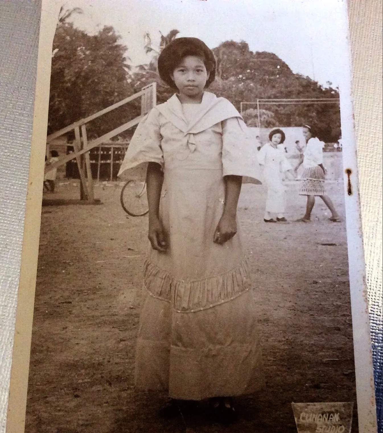 Young girl in light-colored dress with draped collar and fringe hem, holding a badminton racket, standing in an outdoor courtyard with other children in background