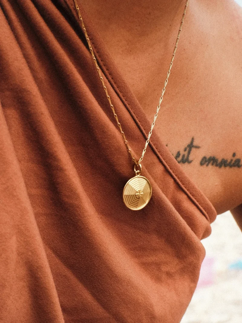 Gold pendant necklace with geometric oval design worn by person in terracotta fabric with visible tattoo