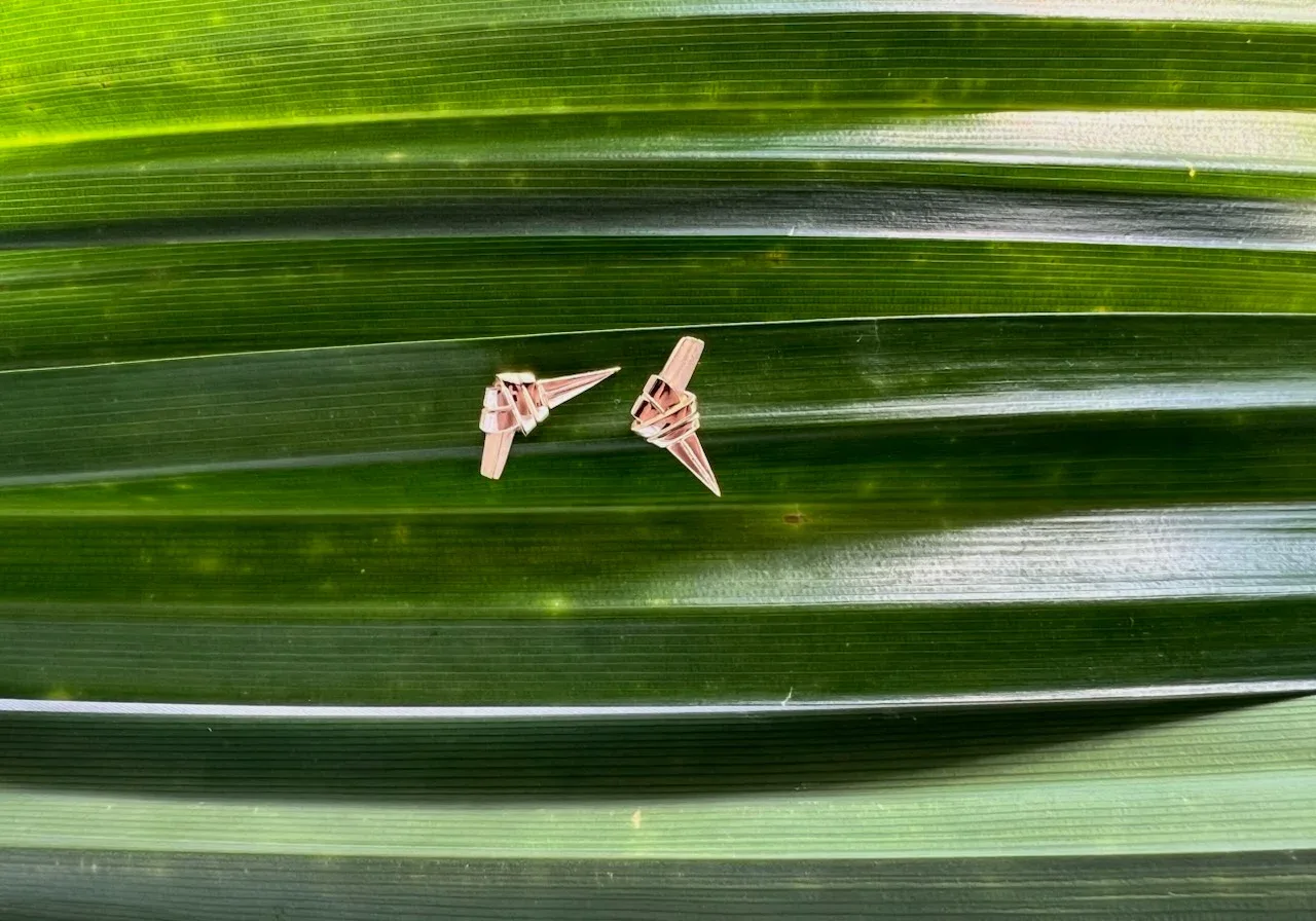 Two small, intricately designed pandan knot gold earrings on a background of layered green leaves.