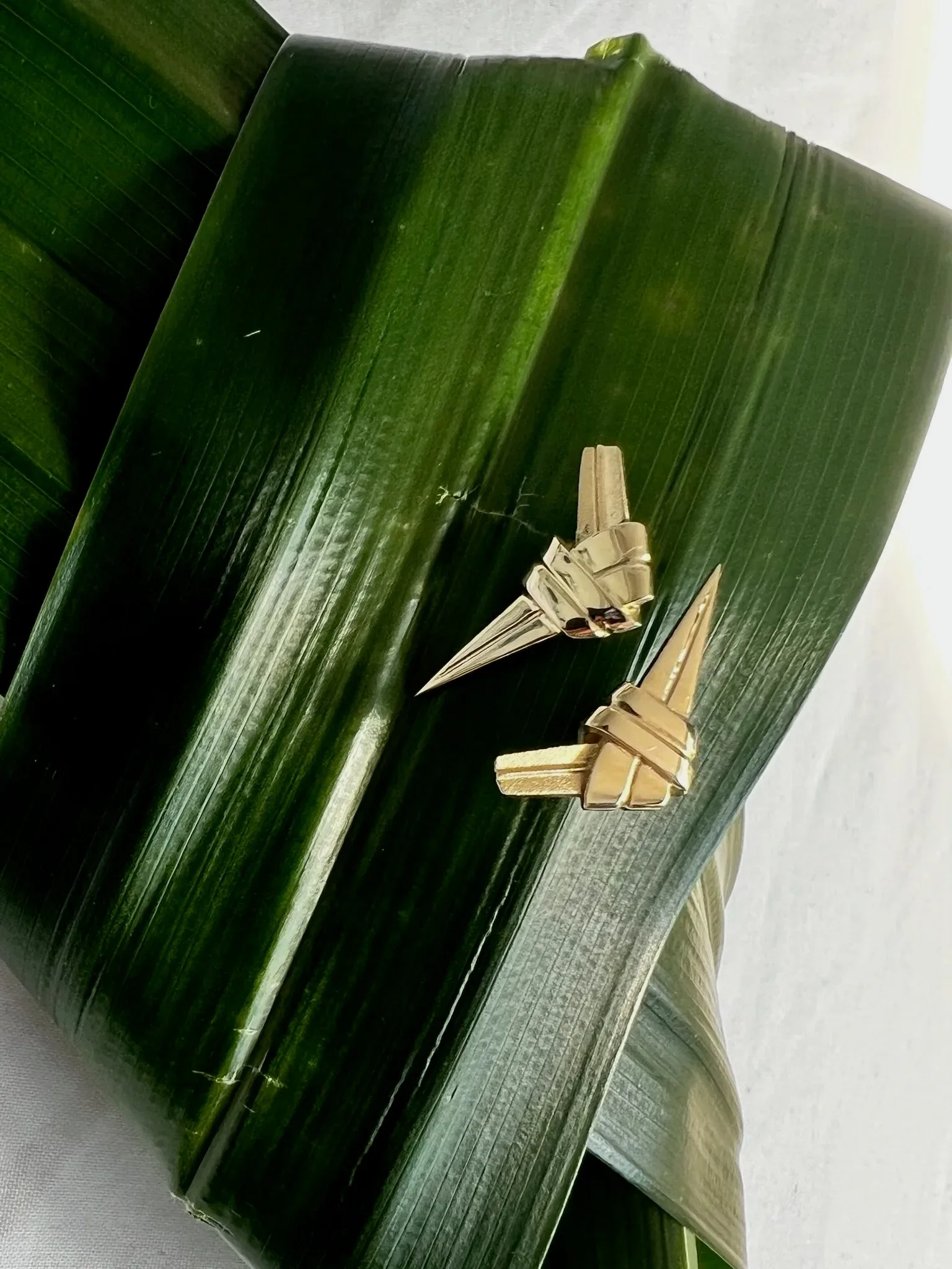 Two gold ear studs with geometric wrapped designs displayed on green banana leaves