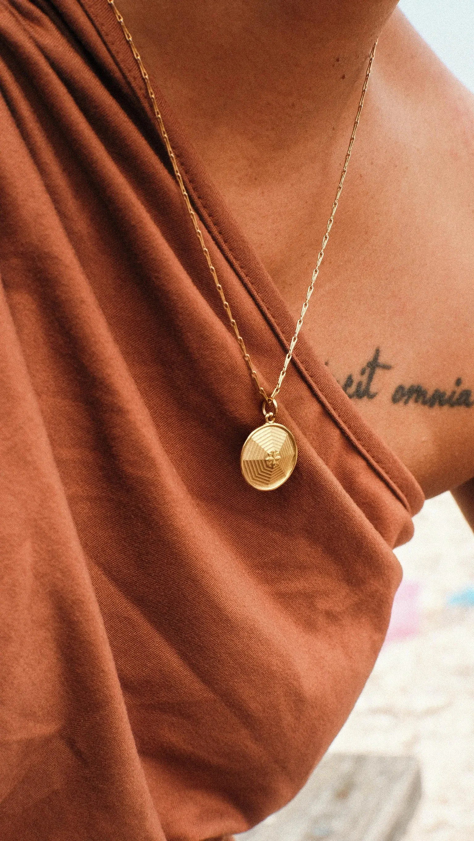 Gold pendant necklace worn on person in rust-colored fabric, showing circular textured design