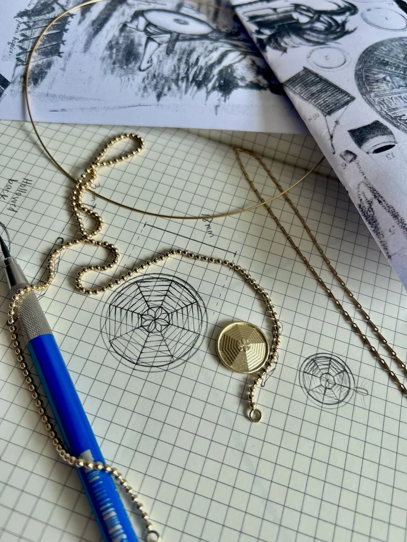 Gold beaded necklace with circular pendant displayed on graph paper with sketches, blue pen, and design references