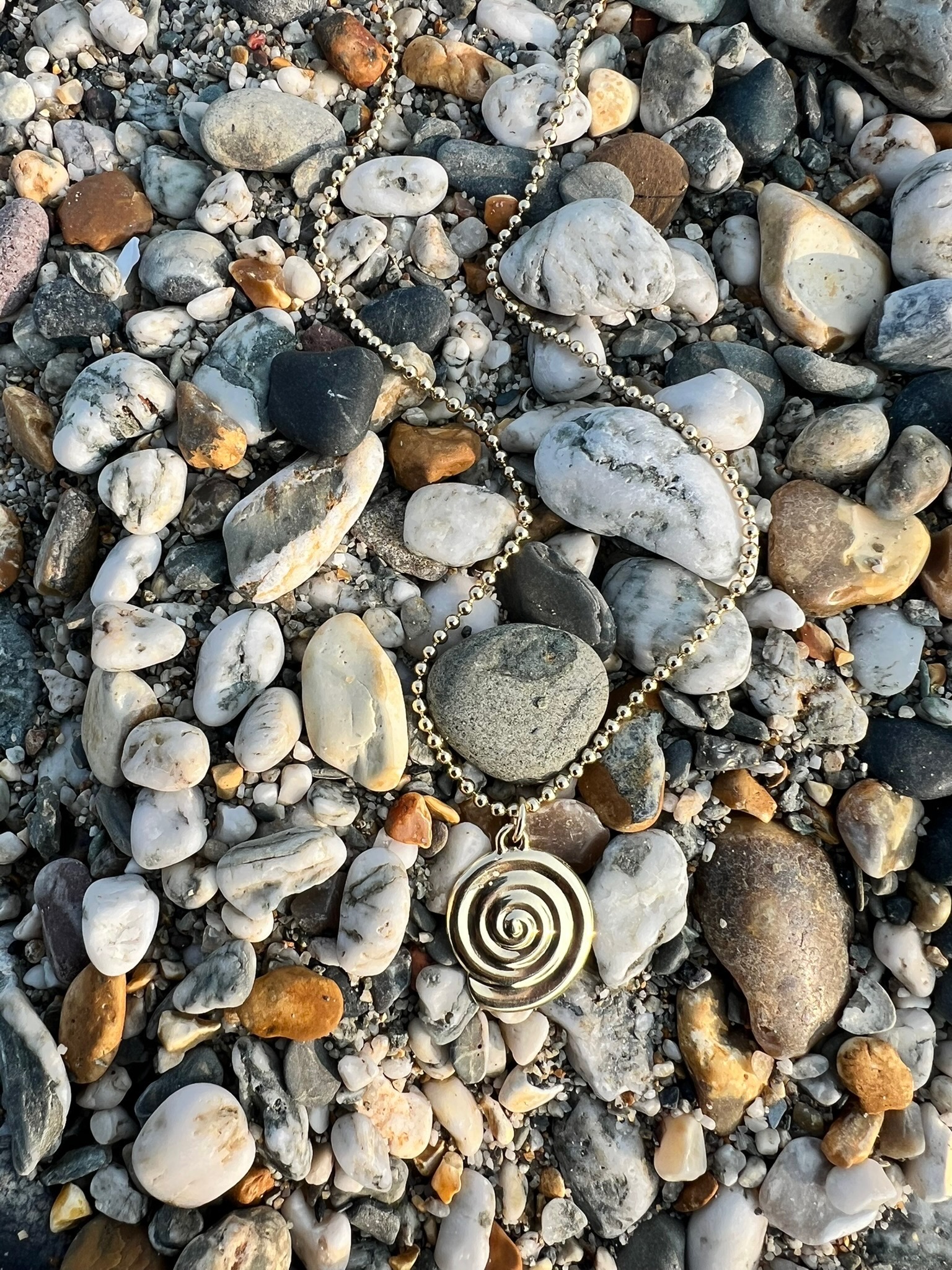 Spiral pendant on a rocky beach, inspired by puka shells, crafted in 9ct yellow gold on a delicate bead chain.