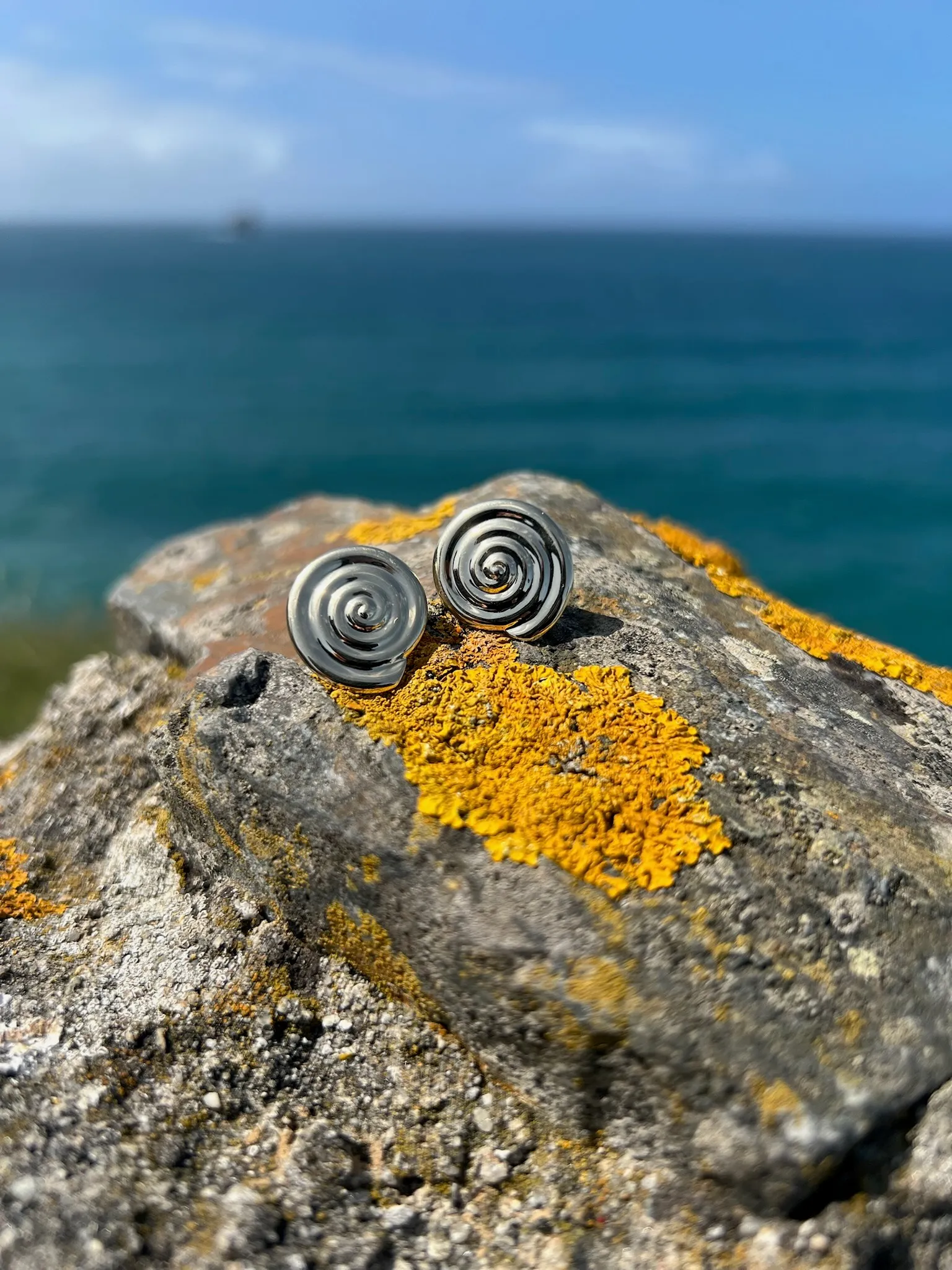 A pair of gold spiral stud earrings resting on a rock, inspired by puka shells from Hawai’i and the Philippines.