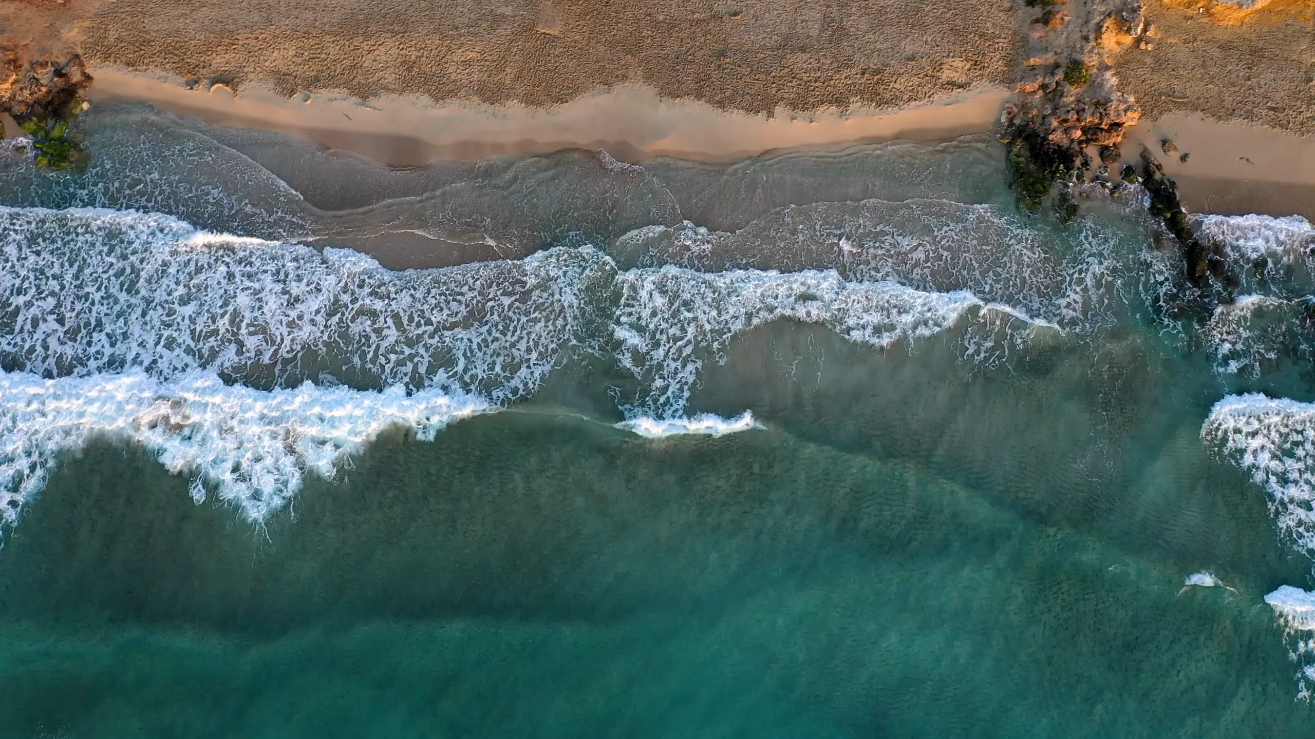 Top-down aerial view of waves crashing on a sandy shoreline with clear, turquoise water. The waves create white foam patterns as they approach the beach, with rocks visible along the edge of the shore and patches of sand meeting coastal vegetation.